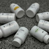 Mehrere weiße Behälter mit Nahrungsergänzungsmitteln liegen auf einer grauen Oberfläche. Etiketten mit verschiedenen Nährstoffen sind sichtbar.