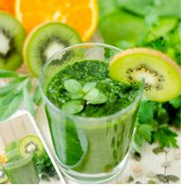 Ein Glas grüner Smoothie, dekoriert mit einer Scheibe Kiwi, steht auf einem Tisch. Im Hintergrund sind frische Zutaten wie Kiwi, Orange und Kräuter zu sehen.